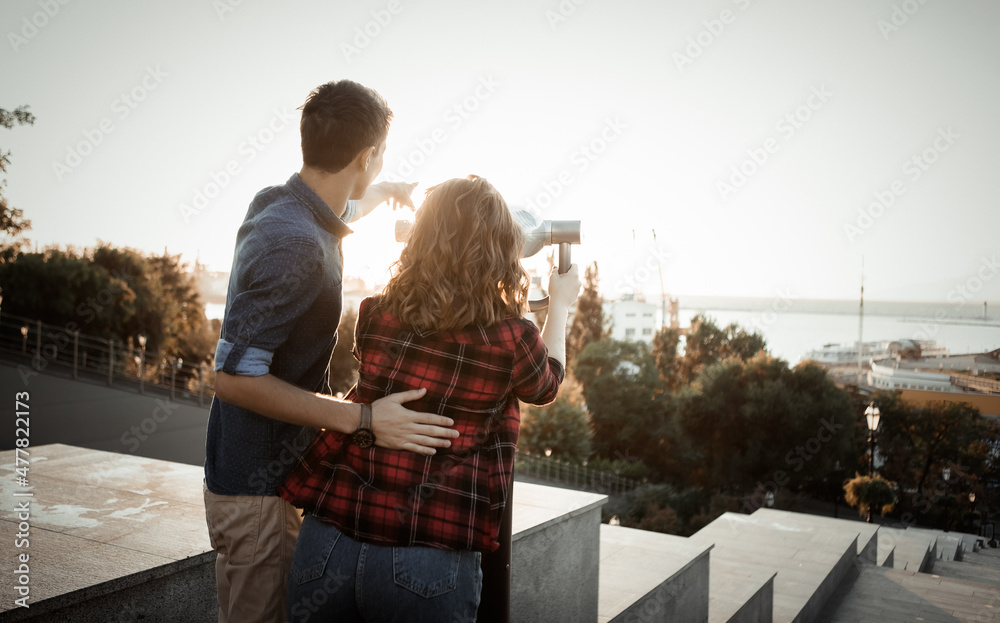Naklejka premium Young couple of tourists look through city binoculars in the city at sunrise. Travel, vacation concept