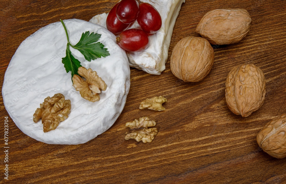 A circle of camembert cheese with white noble mold lies on a wooden board. A piece of cheese with walnuts and grapes.