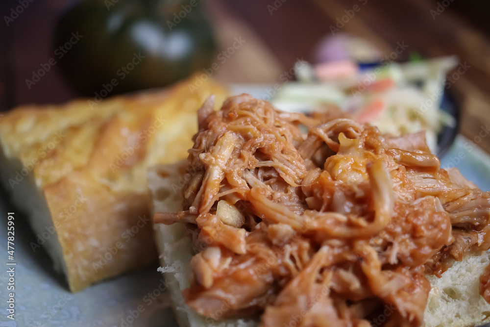 Closeup of dish with marinated pulled pork on fresh baguette, blurred cole slaw salad background