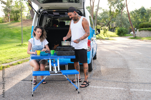 Young tattooed couple cooking in a camping gas kitchen near by the van.