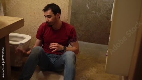 Young bearded drunk man is sitting near the toilet holding his belly and drinking pure water. A guy is suffering from stomach ache after alcohol poisoning. He is feeling queasiness because of hangover photo