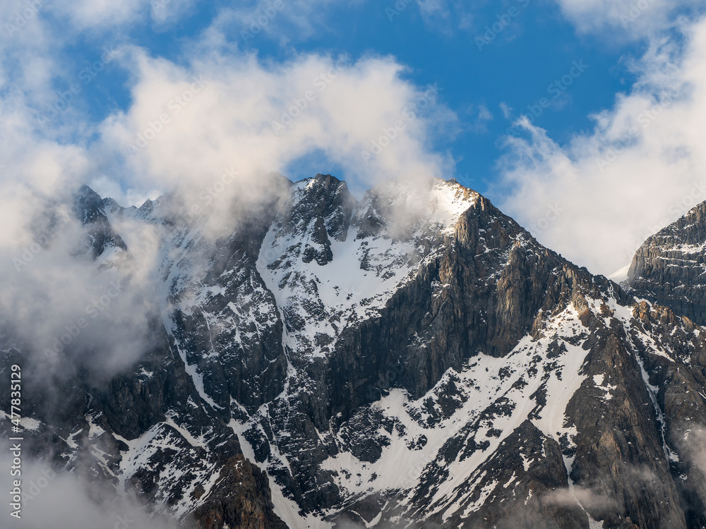 White clouds over a high pointed mountain. Snowy high-altitude alpine landscape with snow-capped mountain peak and sharp rocks under cloudy blue sky. White clouds over a high pointed mountain. Snowy high-altitude alpine landscape with snow-capped mountain peak and sharp rocks under cloudy blue sky.