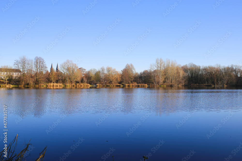 View over lake on bare trees against blue clear winter sky and church - Nettetal (Lobberich), Germany