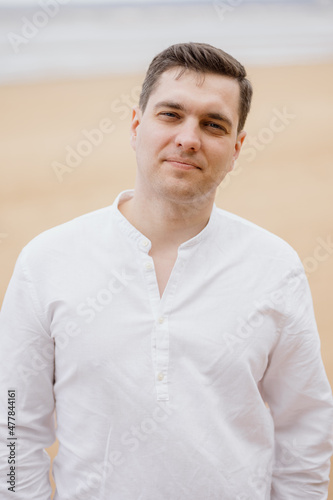 a confident handsome European man with a white shirt walks along the seashore or ocean and smiles looking into the frame. a millennial man is resting in nature and rejoicing. a successful man on