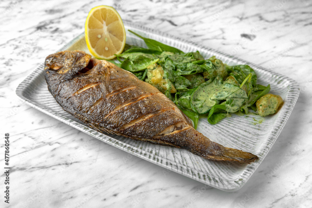 Dorado grilled fish with spinach salad and cheese in a festive plate on a marble background. Restaurant banquet menu. Dorado grilled fish with spinach salad and cheese in a festive plate on a marble background. Restaurant banquet menu.