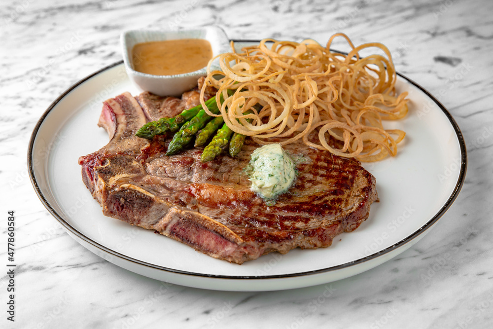 Big delicious juicy bone steak with onion rings in a festive plate on a marble background. Restaurant banquet menu.