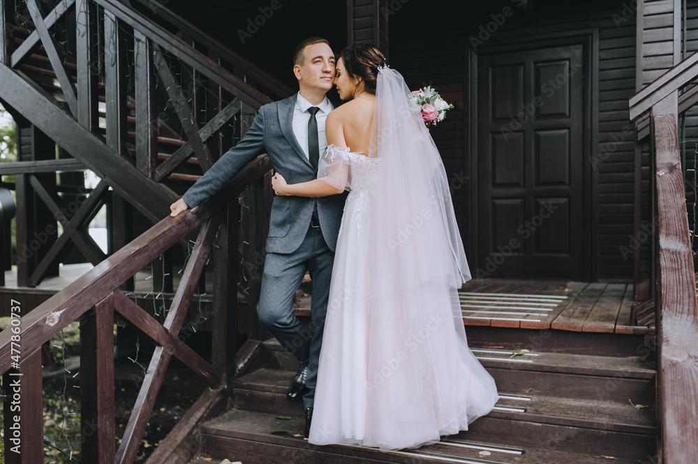 Stylish groom and a beautiful brunette bride in a white dress with a bouquet are standing and hugging on the background of a wooden house with a staircase. Wedding portrait of the newlyweds.