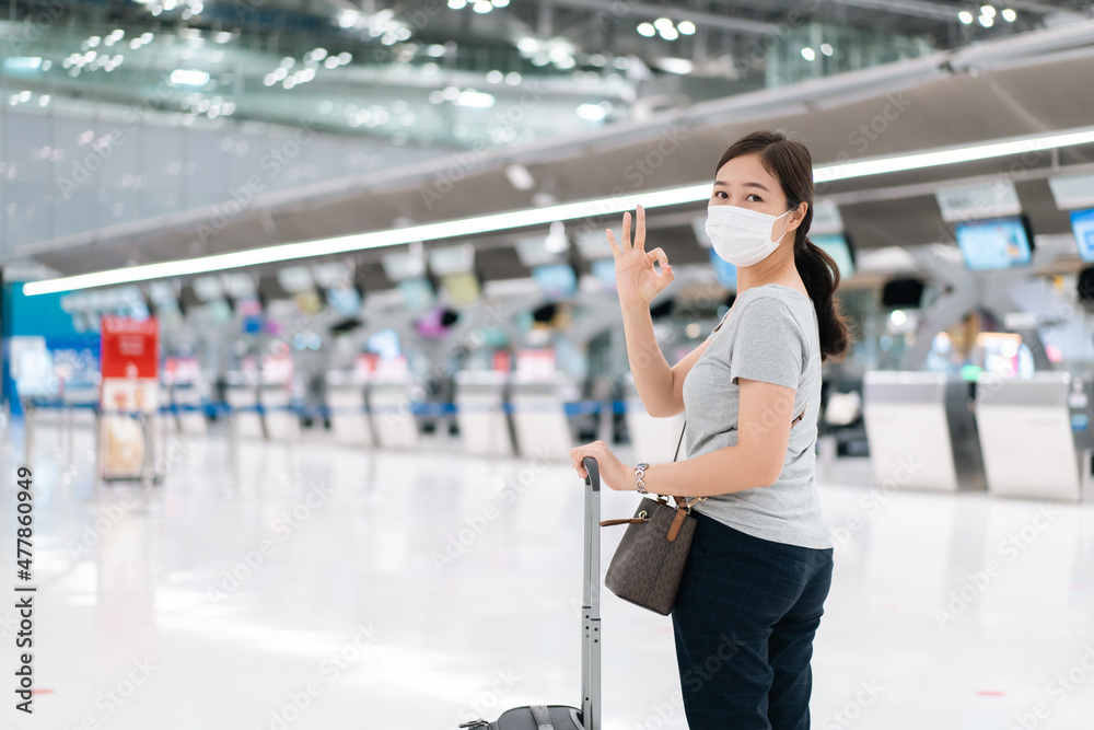 Beautiful Asian young woman with protective face mask looking and smiling to the camera while walking in the airport. Travel and tourist concept during the coronavirus or COVID-19 pandemic.