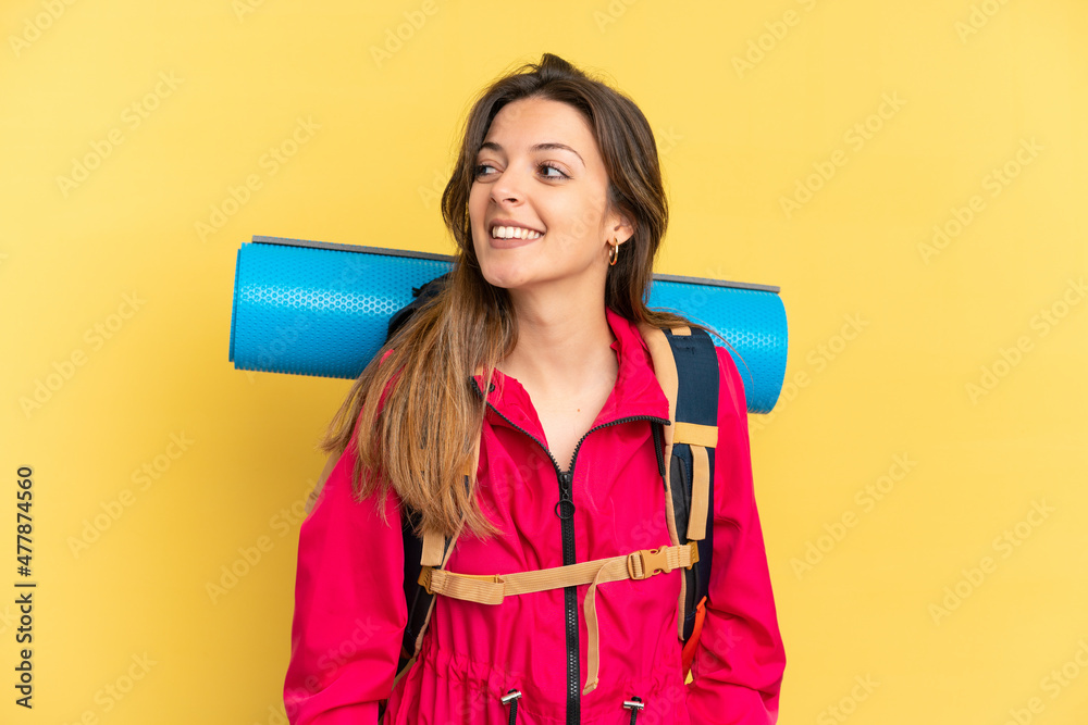 Young mountaineer man with a big backpack isolated on yellow background looking side Young mountaineer man with a big backpack isolated on yellow background looking side