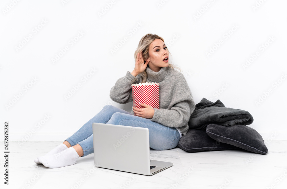 Teenager blonde girl eating popcorn while watching a movie on the laptop listening to something by putting hand on the ear Teenager blonde girl eating popcorn while watching a movie on the laptop listening to something by putting hand on the ear