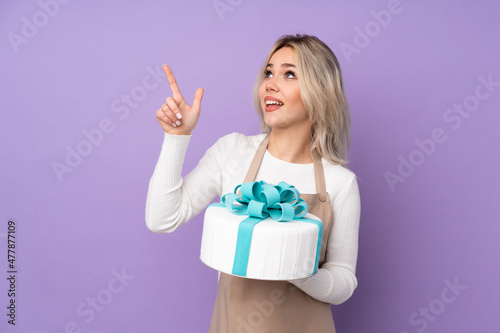 Young pastry chef holding a big cake over isolated purple background pointing with the index finger a great idea