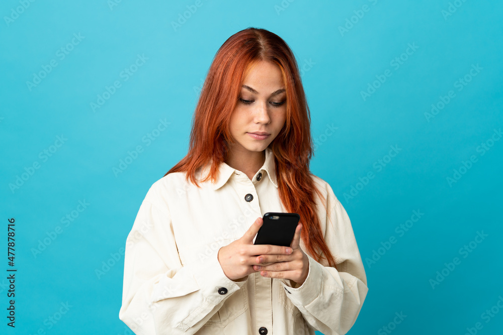 Teenager Russian girl isolated on blue background using mobile phone Teenager Russian girl isolated on blue background using mobile phone