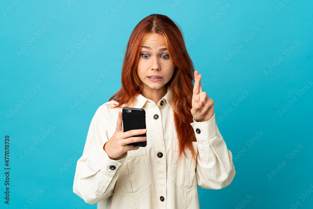 Teenager Russian girl isolated on blue background using mobile phone with fingers crossing Teenager Russian girl isolated on blue background using mobile phone with fingers crossing