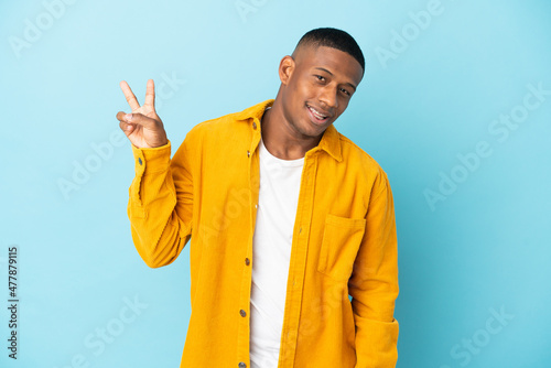 Young latin man isolated on blue background smiling and showing victory sign