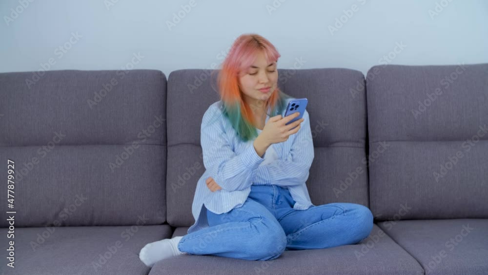 Cute young Caucasian girl with dyed hair using mobile phone for communication while sitting on couch at home on lockdown. Beautiful white woman in late 20s browsing smartphone with smile