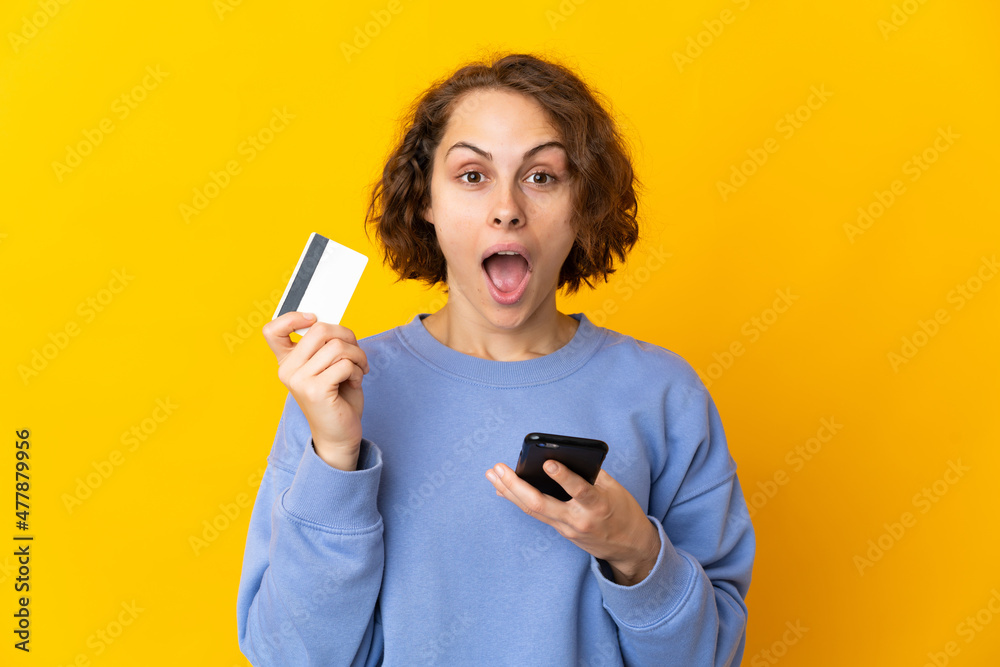 Young English woman isolated on pink background buying with the mobile and holding a credit card with surprised expression