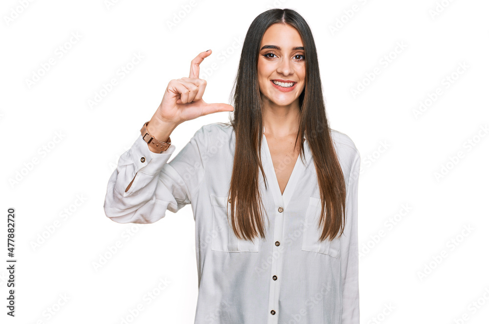Young beautiful woman wearing casual white shirt smiling and confident gesturing with hand doing small size sign with fingers looking and the camera. measure concept. Young beautiful woman wearing casual white shirt smiling and confident gesturing with hand doing small size sign with fingers looking and the camera. measure concept.