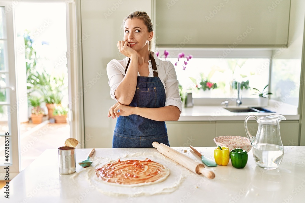 Beautiful blonde woman wearing apron cooking pizza looking stressed and nervous with hands on mouth biting nails. anxiety problem.