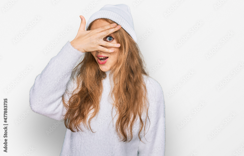 Young caucasian girl wearing wool sweater and winter cap peeking in shock covering face and eyes with hand, looking through fingers with embarrassed expression. Young caucasian girl wearing wool sweater and winter cap peeking in shock covering face and eyes with hand, looking through fingers with embarrassed expression.