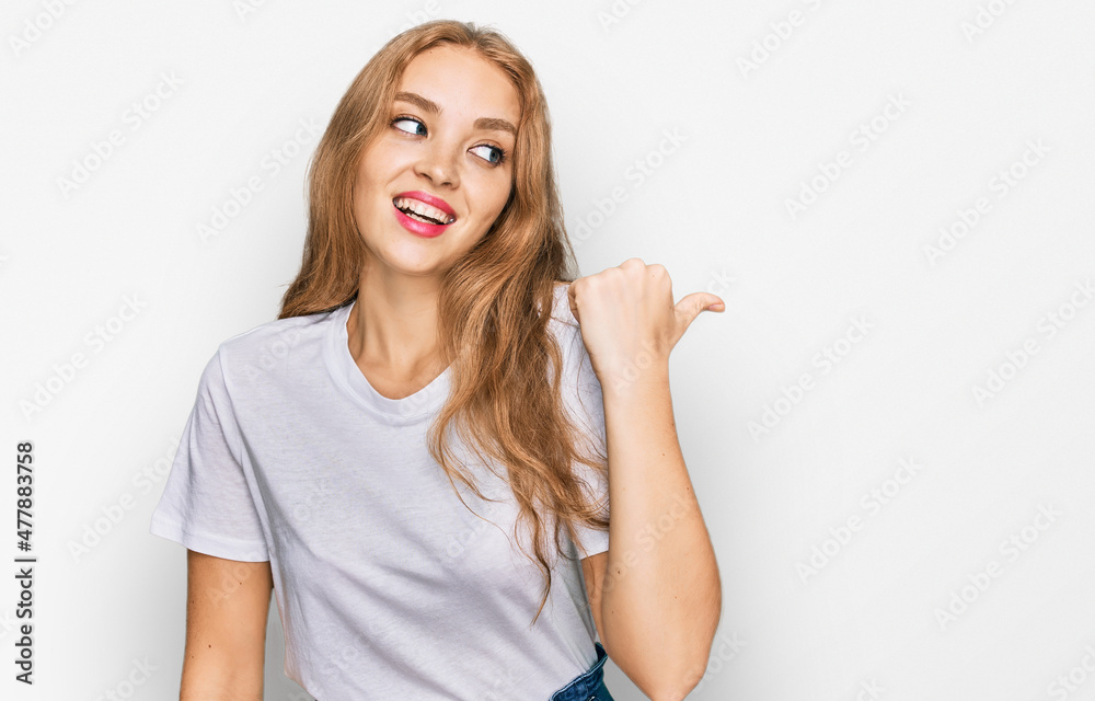 Young caucasian girl wearing casual white t shirt smiling with happy face looking and pointing to the side with thumb up. Young caucasian girl wearing casual white t shirt smiling with happy face looking and pointing to the side with thumb up.