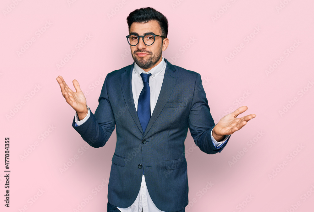 Hispanic man with beard wearing business suit and tie clueless and confused expression with arms and hands raised. doubt concept. Hispanic man with beard wearing business suit and tie clueless and confused expression with arms and hands raised. doubt concept.