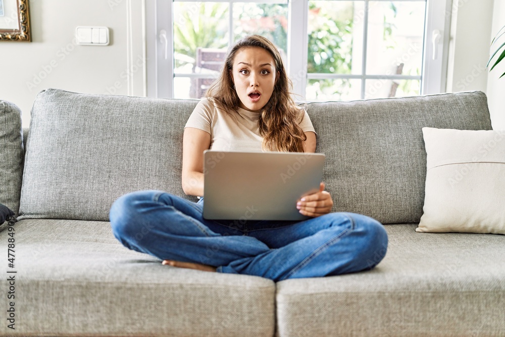 Beautiful young brunette woman sitting on the sofa using computer laptop at home afraid and shocked with surprise expression, fear and excited face. Beautiful young brunette woman sitting on the sofa using computer laptop at home afraid and shocked with surprise expression, fear and excited face.