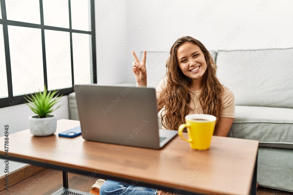 Beautiful hispanic woman using computer laptop at home smiling with happy face winking at the camera doing victory sign. number two. Beautiful hispanic woman using computer laptop at home smiling with happy face winking at the camera doing victory sign. number two.
