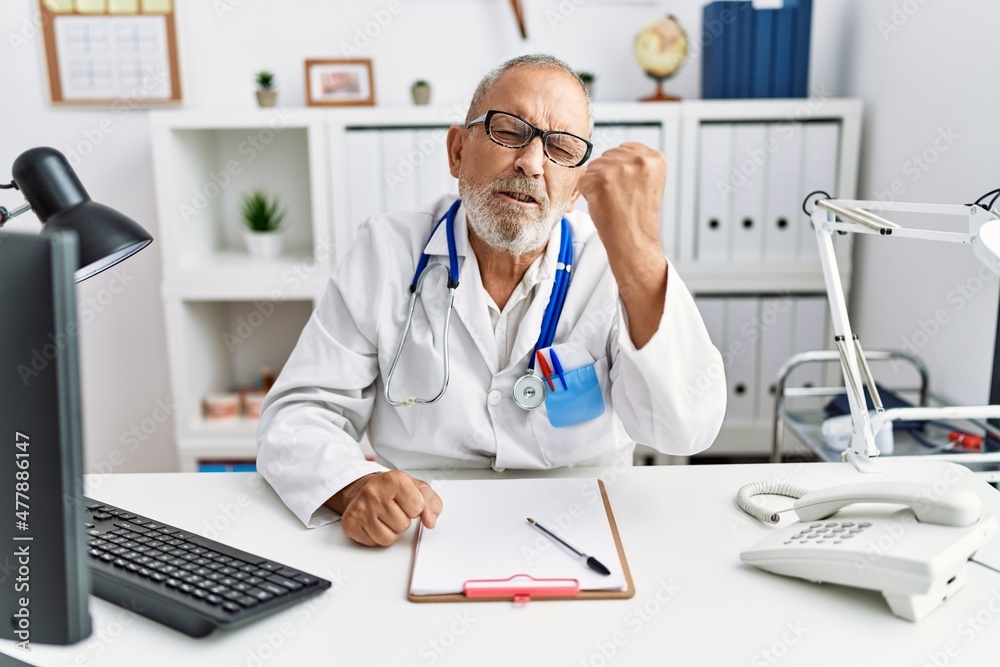 Mature doctor man at the clinic angry and mad raising fist frustrated and furious while shouting with anger. rage and aggressive concept.