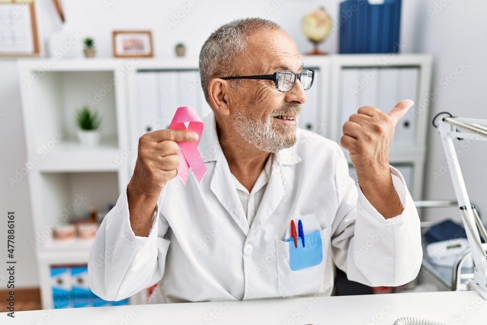 Mature doctor man holding pink cancer ribbon at the clinic pointing thumb up to the side smiling happy with open mouth Mature doctor man holding pink cancer ribbon at the clinic pointing thumb up to the side smiling happy with open mouth