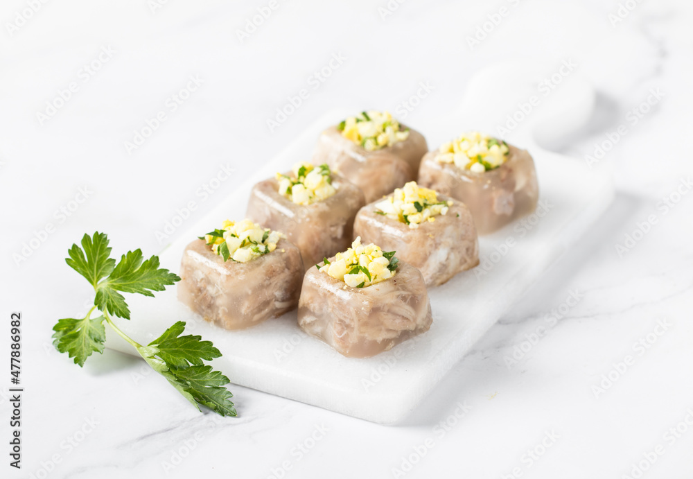 Cold appetizer. Pork jelly with pieces of meat in the form of small square portions with a filling of finely chopped egg and parsley, on a serving board on a white background