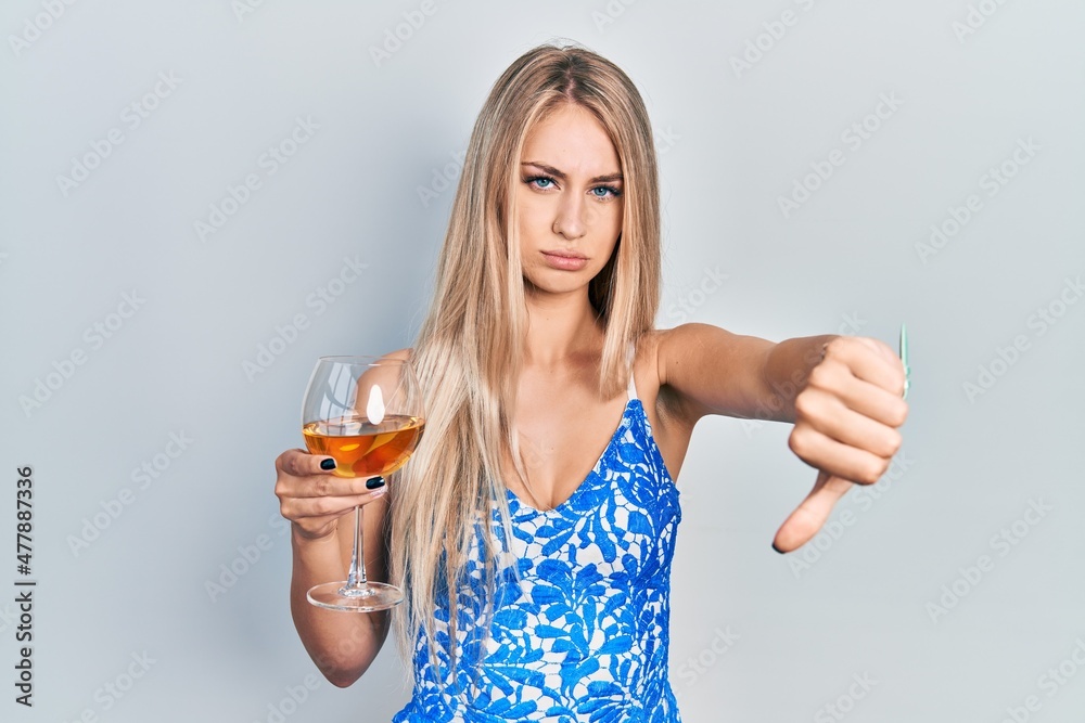 Young beautiful caucasian woman drinking a glass of white wine looking unhappy and angry showing rejection and negative with thumbs down gesture. bad expression. Young beautiful caucasian woman drinking a glass of white wine looking unhappy and angry showing rejection and negative with thumbs down gesture. bad expression.