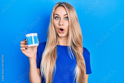Young caucasian woman holding earwax cotton removers scared and amazed with open mouth for surprise, disbelief face photo