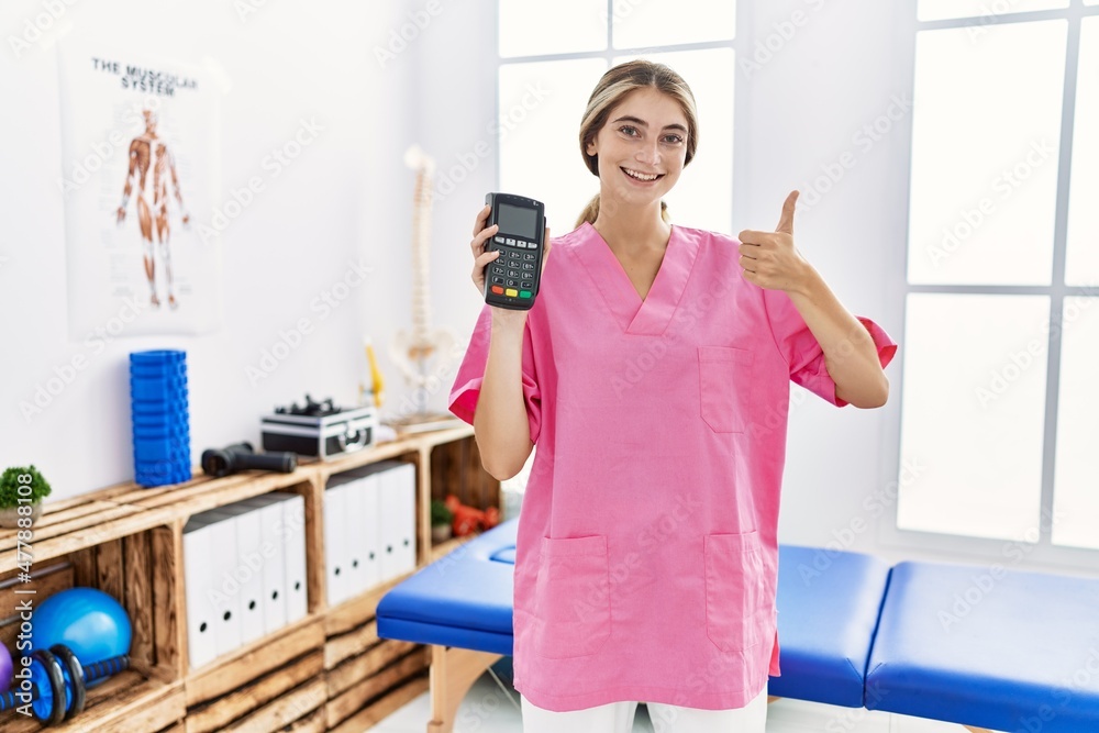 Young physiotherapist woman working at pain recovery clinic holding dataphone smiling happy and positive, thumb up doing excellent and approval sign Young physiotherapist woman working at pain recovery clinic holding dataphone smiling happy and positive, thumb up doing excellent and approval sign