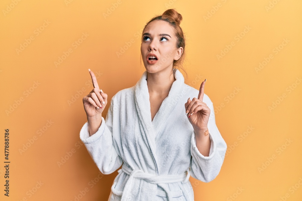 Young blonde woman wearing bathrobe amazed and surprised looking up and pointing with fingers and raised arms. Young blonde woman wearing bathrobe amazed and surprised looking up and pointing with fingers and raised arms.