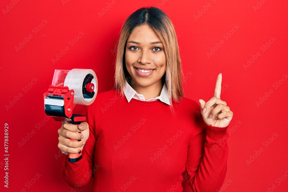 Beautiful hispanic woman holding packing tape smiling with an idea or question pointing finger with happy face, number one Beautiful hispanic woman holding packing tape smiling with an idea or question pointing finger with happy face, number one