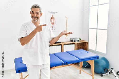 Middle age hispanic therapist man working at pain recovery clinic amazed and smiling to the camera while presenting with hand and pointing with finger.