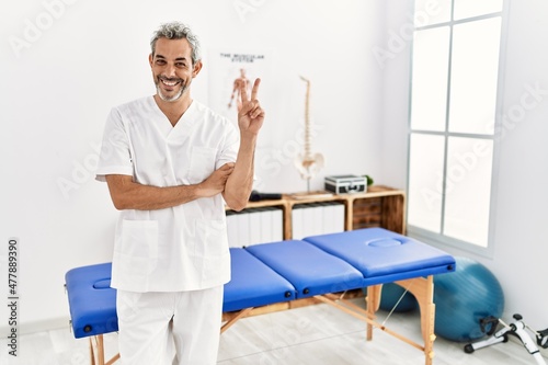 Middle age hispanic therapist man working at pain recovery clinic smiling with happy face winking at the camera doing victory sign with fingers. number two.