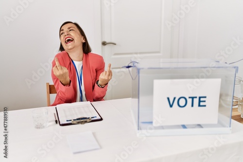 Beautiful middle age hispanic woman at political election sitting by ballot showing middle finger doing fuck you bad expression, provocation and rude attitude. screaming excited