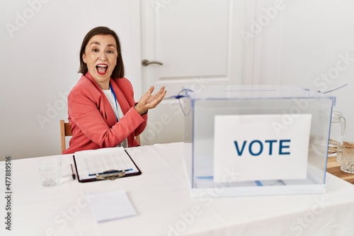 Beautiful middle age hispanic woman at political election sitting by ballot pointing aside with hands open palms showing copy space, presenting advertisement smiling excited happy