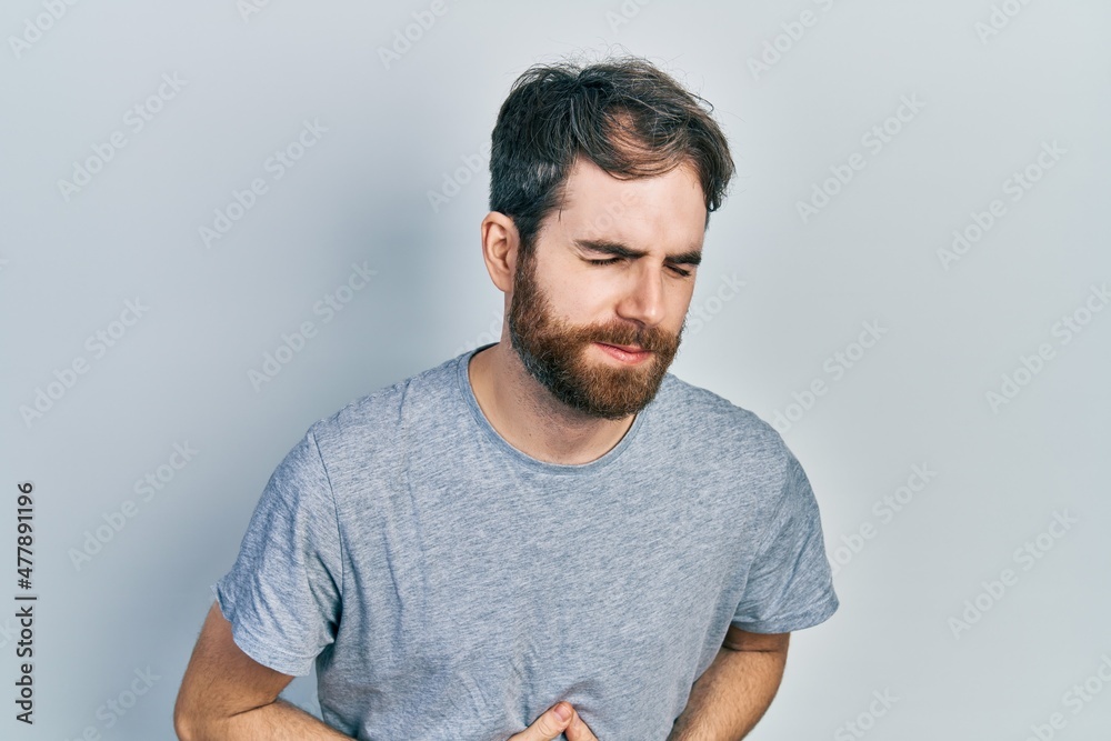 Caucasian man with beard wearing casual grey t shirt with hand on stomach because indigestion, painful illness feeling unwell. ache concept. Caucasian man with beard wearing casual grey t shirt with hand on stomach because indigestion, painful illness feeling unwell. ache concept.