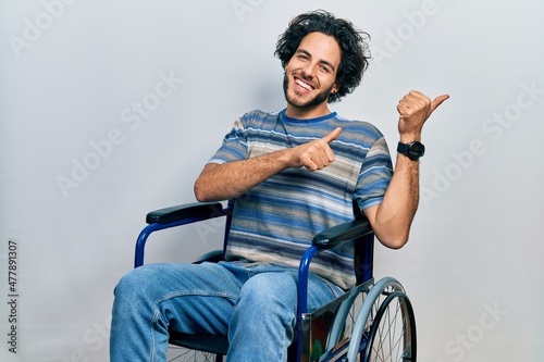 Handsome hispanic man sitting on wheelchair pointing to the back behind with hand and thumbs up, smiling confident