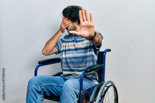 Handsome hispanic man sitting on wheelchair covering eyes with hands and doing stop gesture with sad and fear expression. embarrassed and negative concept.
