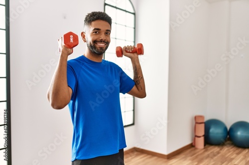 Handsome hispanic man doing exercise training at the gym holding dumbbells