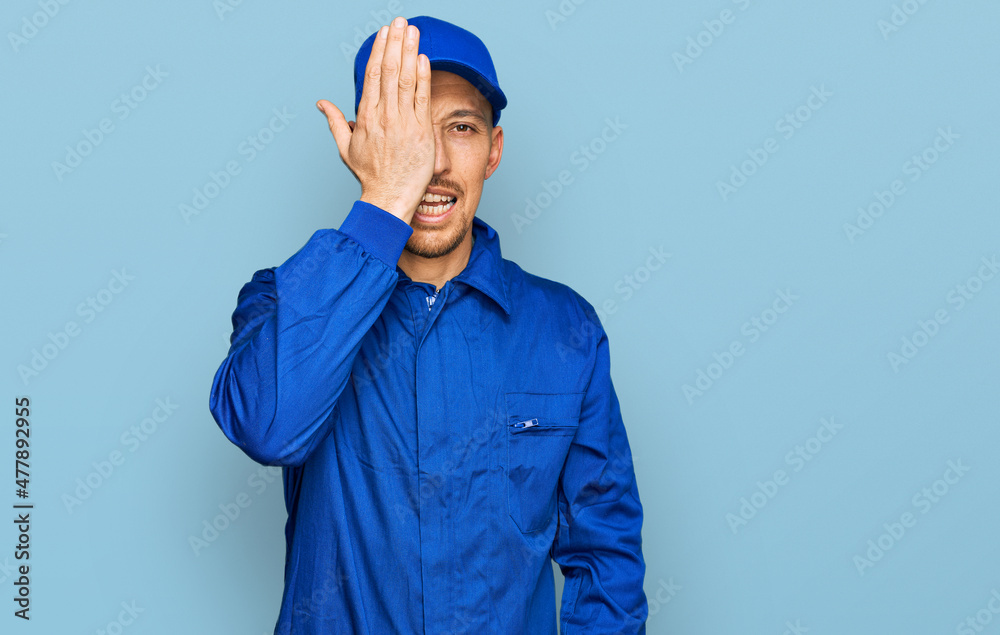 Bald man with beard wearing builder jumpsuit uniform covering one eye with hand, confident smile on face and surprise emotion. Bald man with beard wearing builder jumpsuit uniform covering one eye with hand, confident smile on face and surprise emotion.