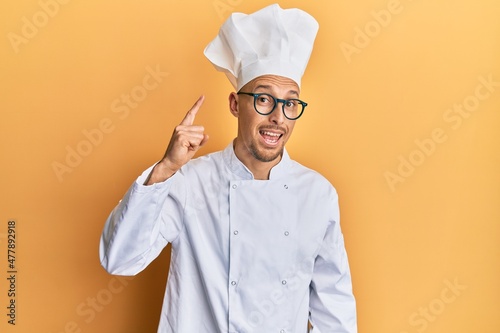 Bald man with beard wearing professional cook uniform pointing finger up with successful idea. exited and happy. number one.