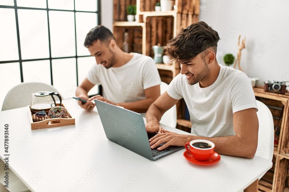 Obraz premium Two hispanic men couple having breakfast using smartphone and laptop at home