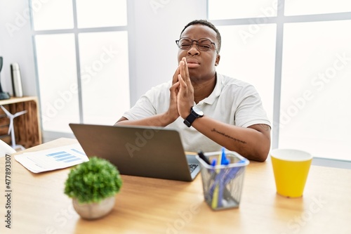 Young african man working at the office using computer laptop begging and praying with hands together with hope expression on face very emotional and worried. begging.