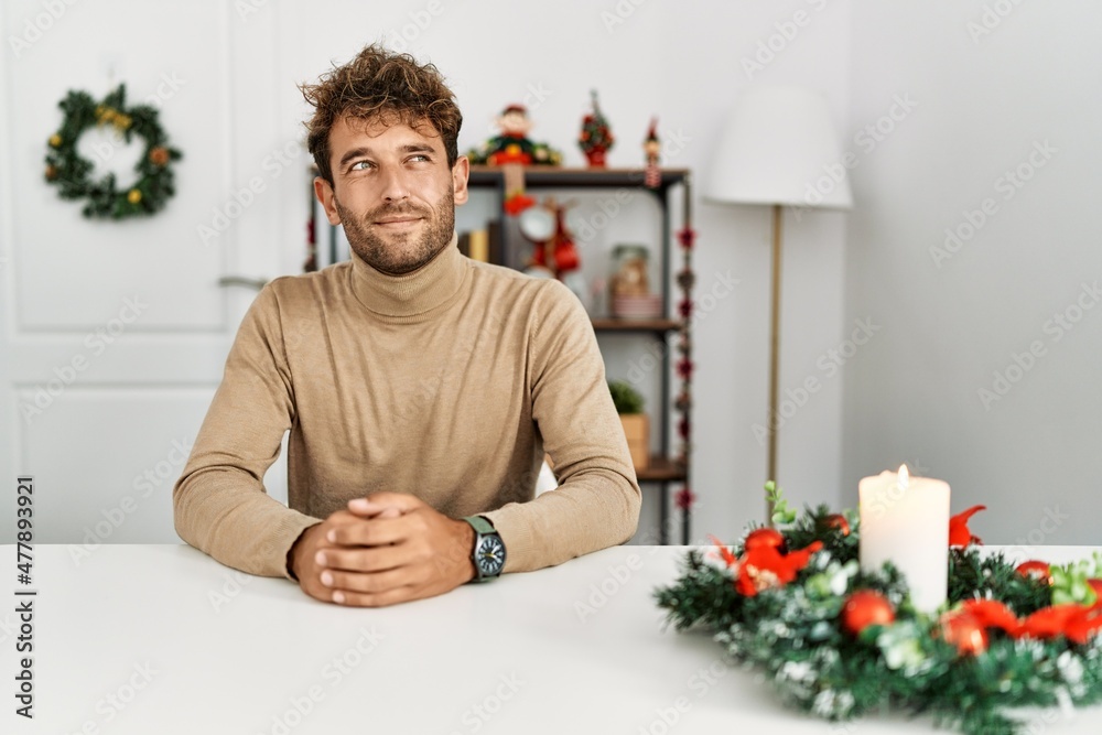 Young handsome man with beard sitting on the table by christmas decoration smiling looking to the side and staring away thinking.