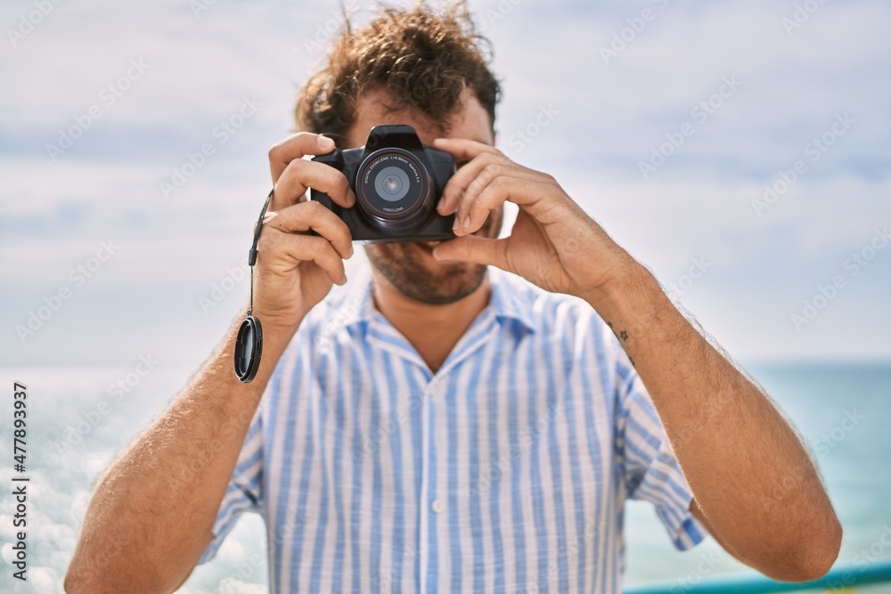 Obraz premium Young hispanic man smiling happy using camera at the beach.