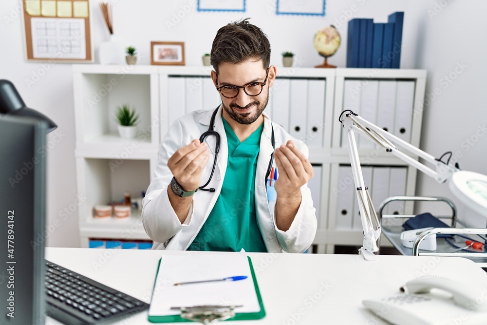 Young man with beard wearing doctor uniform and stethoscope at the clinic doing money gesture with hands, asking for salary payment, millionaire business Young man with beard wearing doctor uniform and stethoscope at the clinic doing money gesture with hands, asking for salary payment, millionaire business